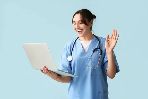 Beautiful female doctor with laptop waving hand on blue background