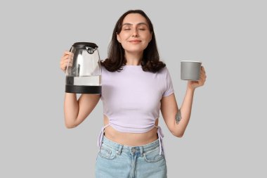 Gri arka planda modern elektrikli su ısıtıcısı ve kupası olan rüya gören genç kadın.