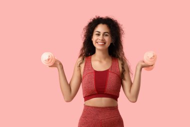 Spor kıyafetli genç bir Afro-Amerikalı kadın pembe arka planda dambıllarıyla egzersiz yapıyor.