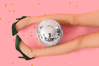 Disko topu ve pembe arka planda konfeti olan şık topuklu kadın bacakları.