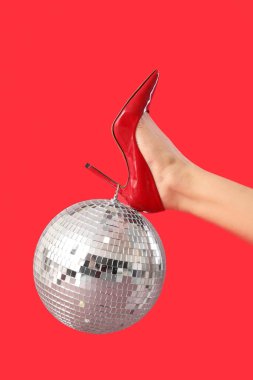 Kırmızı arka planda disko topu olan şık topuklu bir kadın bacağı.
