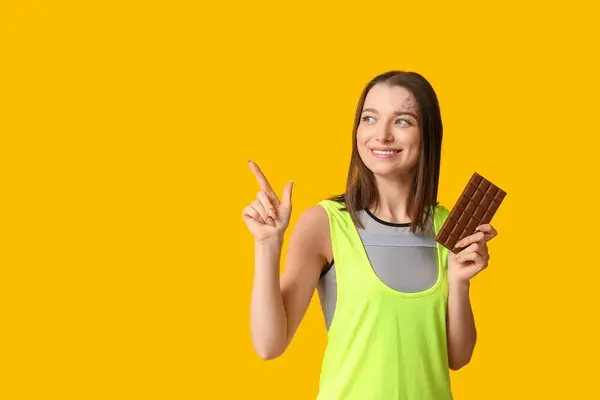 Mladá Žena Drží Čokoládovou Tyčinku Ukazuje Něco Žlutém Pozadí — Stock fotografie