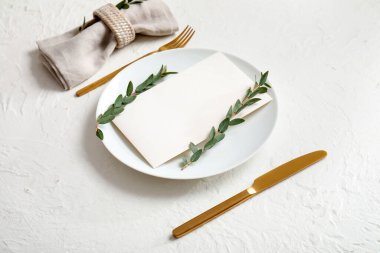 Boş davetiye kartı, çatal bıçak takımı ve beyaz masa üzerinde okaliptüs dallarıyla temiz tabak.