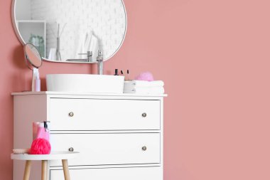 Göğüs çekmecesinde banyo aksesuarları olan beyaz lavabo ve odanın renk duvarının yanındaki masa.