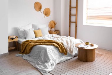 Beyaz battaniyeli rahat bir yatak ve aydınlık yatak odasının içinde bir sehpa.