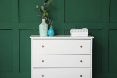 Okaliptüs dallı vazo, yeşil duvarın yanındaki beyaz çekmecelerin üzerinde.