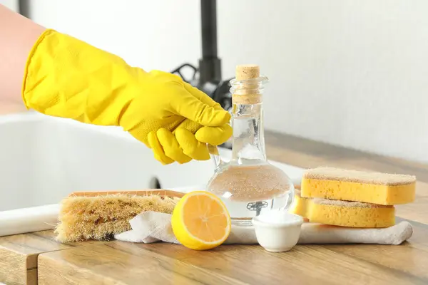 Female hand in rubber glove with jug of vinegar, sponges, brush, baking soda and lemon on wooden table