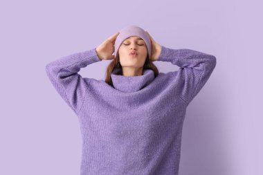 Şapkalı ve örgü süveterli genç bir kadın leylak arka planda öpüşüyor.