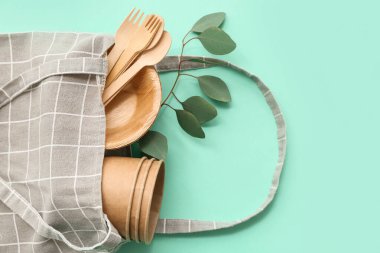 Tek kullanımlık eko masa eşyası ve turkuaz arka planda bitki dalı olan tekstil çantası, yakın plan.