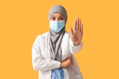 Yüz maskeli kadın Müslüman doktor turuncu arka planda dur hareketi gösteriyor. Dünya Başörtüsü Günü konsepti