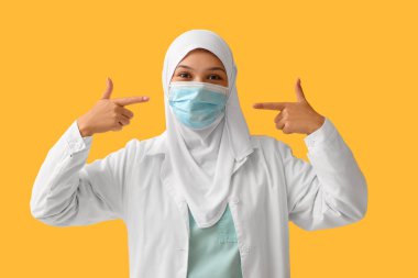 Turuncu arka plandaki yüz maskesine işaret eden tesettürlü kadın Müslüman doktor. Dünya Başörtüsü Günü konsepti