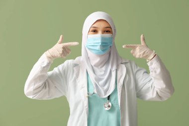 Yeşil arka plandaki yüz maskesine işaret eden tesettürlü kadın Müslüman doktor. Dünya Başörtüsü Günü konsepti