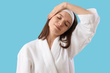 Bornozlu genç bir kadın mavi arka planda yüz geliştirme egzersizi yapıyor.