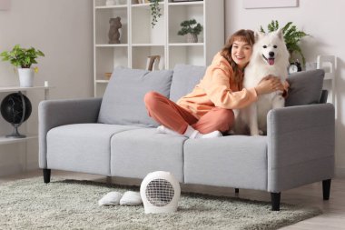 Samoyed köpeğiyle evdeki elektrikli vantilatör ısıtıcısının yanında ısınan genç kadın.