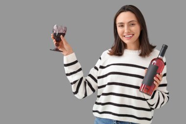 Gri arka planda cam ve kırmızı şarap şişesiyle güzel genç bir kadın.