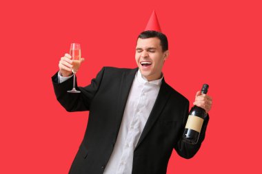 Kırmızı arka planda Noel 'i kutlamak için bir şişe şampanya ve parti şapkalı mutlu genç adam.
