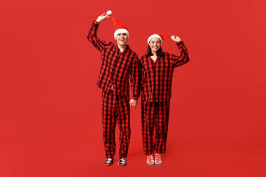 Noel pijamaları ve kırmızı arka planda el ele tutuşan Noel Baba şapkaları giyen mutlu çift.