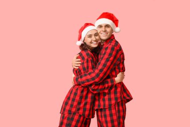 Mutlu genç çift, Noel pijamaları ve Noel Baba şapkaları ile pembe arka plana sarılıyorlar.