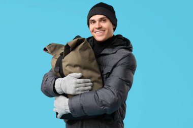 Şık kabarık ceketli ve mavi arka planda çantalı genç bir adam.