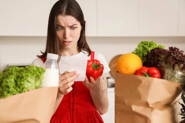 Shocked young woman with bags of food and store receipt in kitchen. Price rise concept