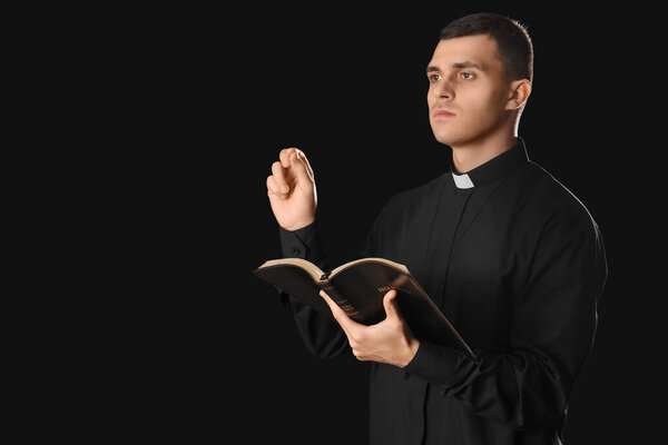 Young priest with Holy Bible on black background
