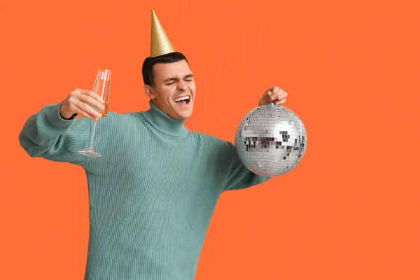 Happy young man in party hat with disco ball and glass of champagne celebrating Christmas on orange background