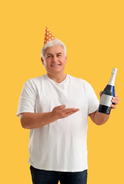 Senior man in party hat with bottle of champagne celebrating Christmas on orange background