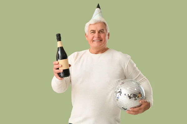 Happy senior man in party hat with bottle of champagne and disco ball celebrating Christmas on green background
