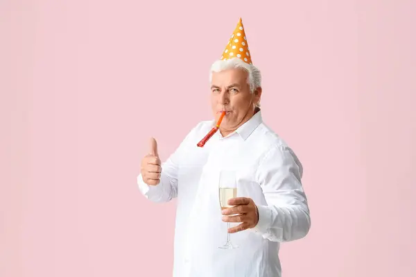 Senior man in party hat with glass of champagne celebrating Christmas and showing thumb-up gesture on pink background
