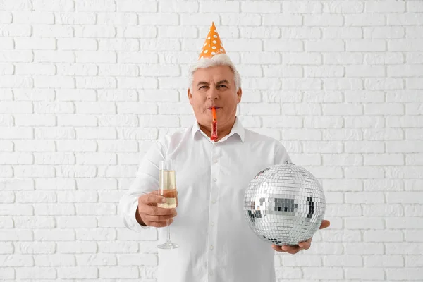 Senior man in party hat with glass of champagne and disco ball celebrating Christmas on white brick background