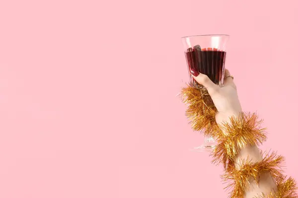 Female hand with glass of warm mulled wine and Christmas decor on pink background