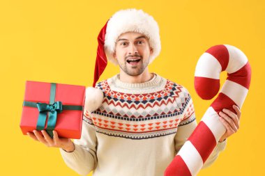 Noel Baba şapkalı yakışıklı adam, büyük şeker kamışı ve sarı arka planda Noel hediyesi.
