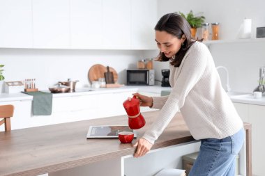 Bir fincan espresso ve gayzer kahve makinesiyle genç ve güzel bir kadın modern mutfakta dergi okuyor.