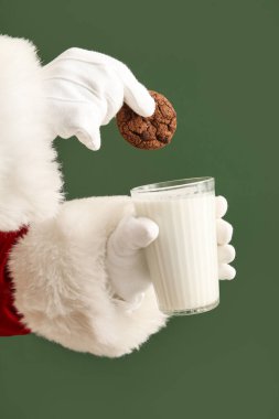 Yeşil arka planda bir bardak süt ve kurabiye ile Noel Baba elleri, yakın plan.