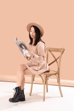Genç Asyalı kadın arka planda sandalyede dergi okuyor.