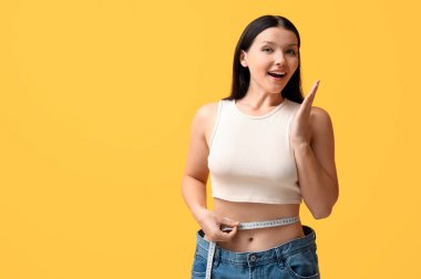 Sarı arka planda ölçüm bandıyla bol kot giymiş güzel, genç bir kadın. Kilo verme konsepti