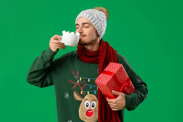 Young man with cocoa and Christmas gift on green background