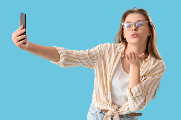Young woman in eyeglasses taking selfie on blue background