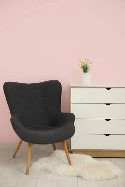 Dark grey armchair with dresser, fluffy rug and daisy near pink wall