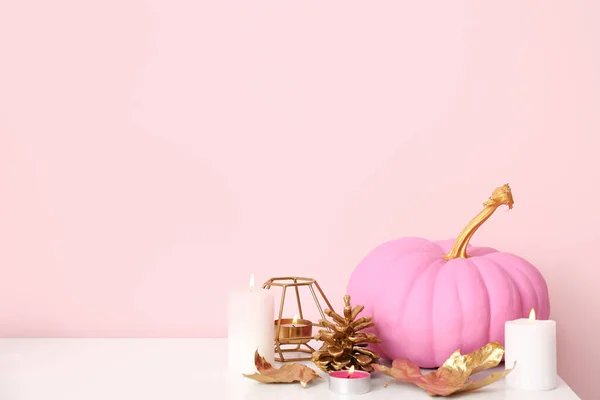 Pink painted pumpkin with autumn leaves and burning candles on white table near color wall