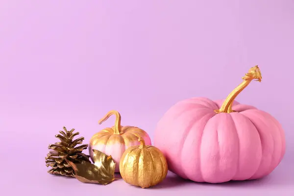 Painted pumpkins with golden leaf and cone on lilac background