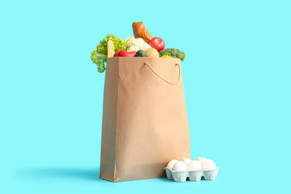 Paper bag with different fresh vegetables and box with eggs on blue background