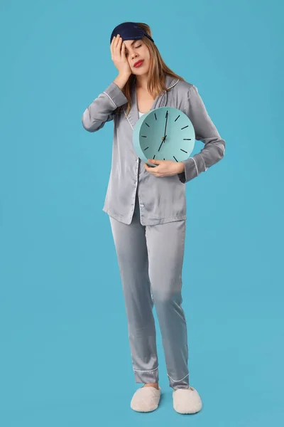 Portrait of tired young woman in pajamas with clock on blue background
