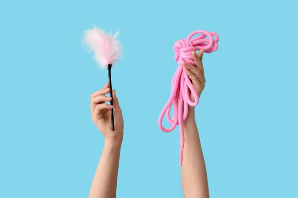 Female hands with feather stick and rope from sex shop on color background, closeup