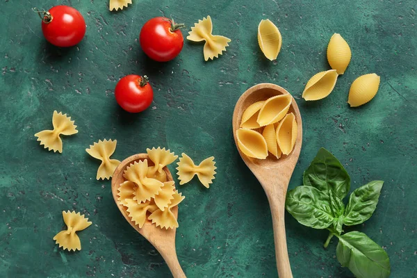 Spoons with different uncooked pasta, basil and tomatoes on green background, closeup