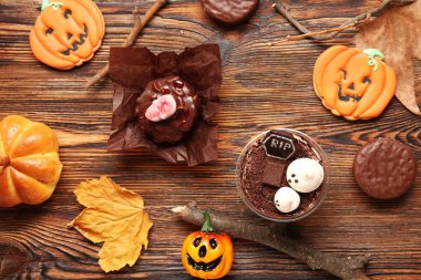 Lezzetli tiramisulu plastik bardak, çikolatalı pasta, kurabiye ve kuru yapraklar Cadılar Bayramı kutlaması için ahşap arka planda.