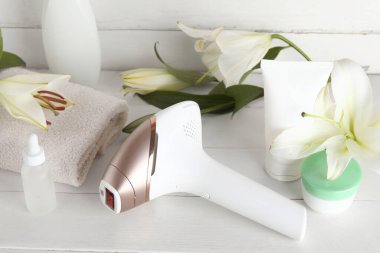 Banyo havlusu, kozmetik ürünleri ve beyaz ahşap arka planda zambak çiçekleri olan modern fotoepilatör.
