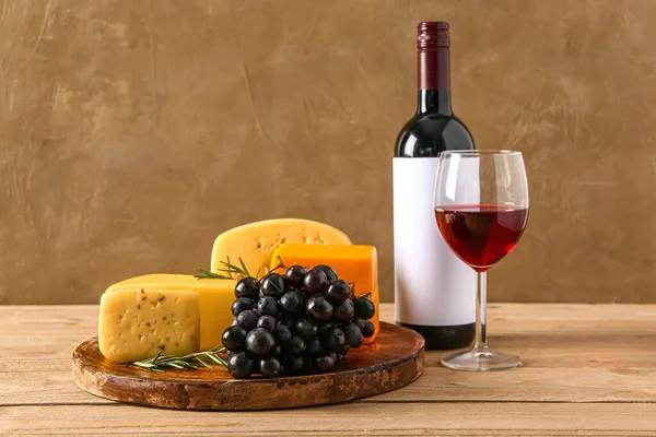 Plate with tasty cheese, grapes, bottle and glass of wine on table near brown wall