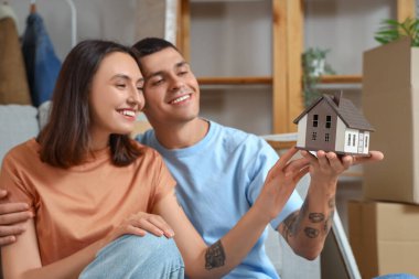 Happy young couple with house model in room on moving day clipart