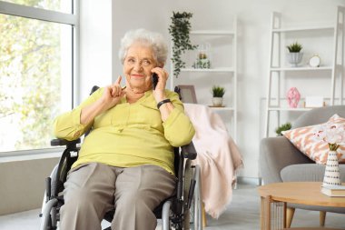 Tekerlekli sandalyedeki yaşlı kadın evde cep telefonuyla konuşuyor.